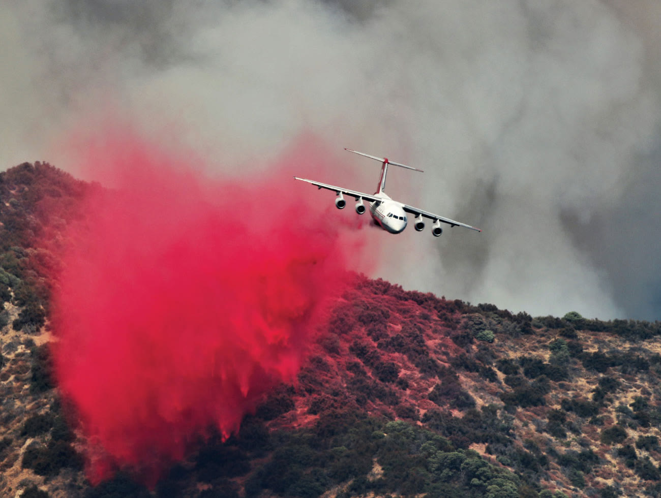 Four-engine aerial tanker releasing a large red fire-retardant cloud over a smoky hillside