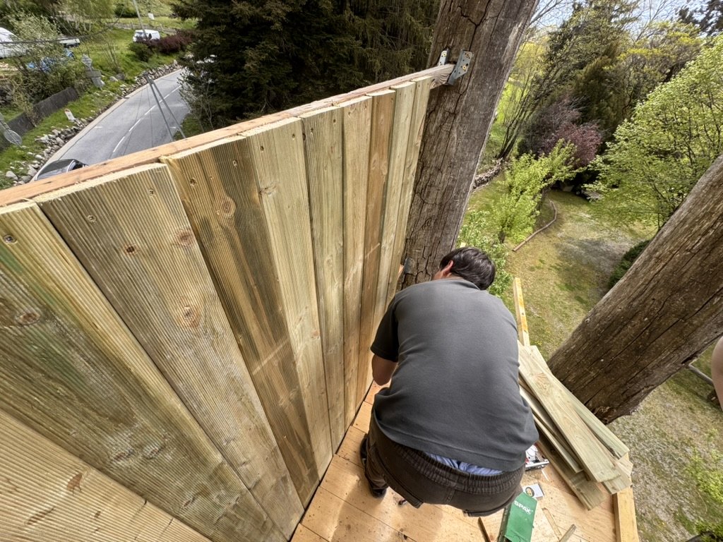 Person fitting vertical timber boards on an elevated platform between two trunks