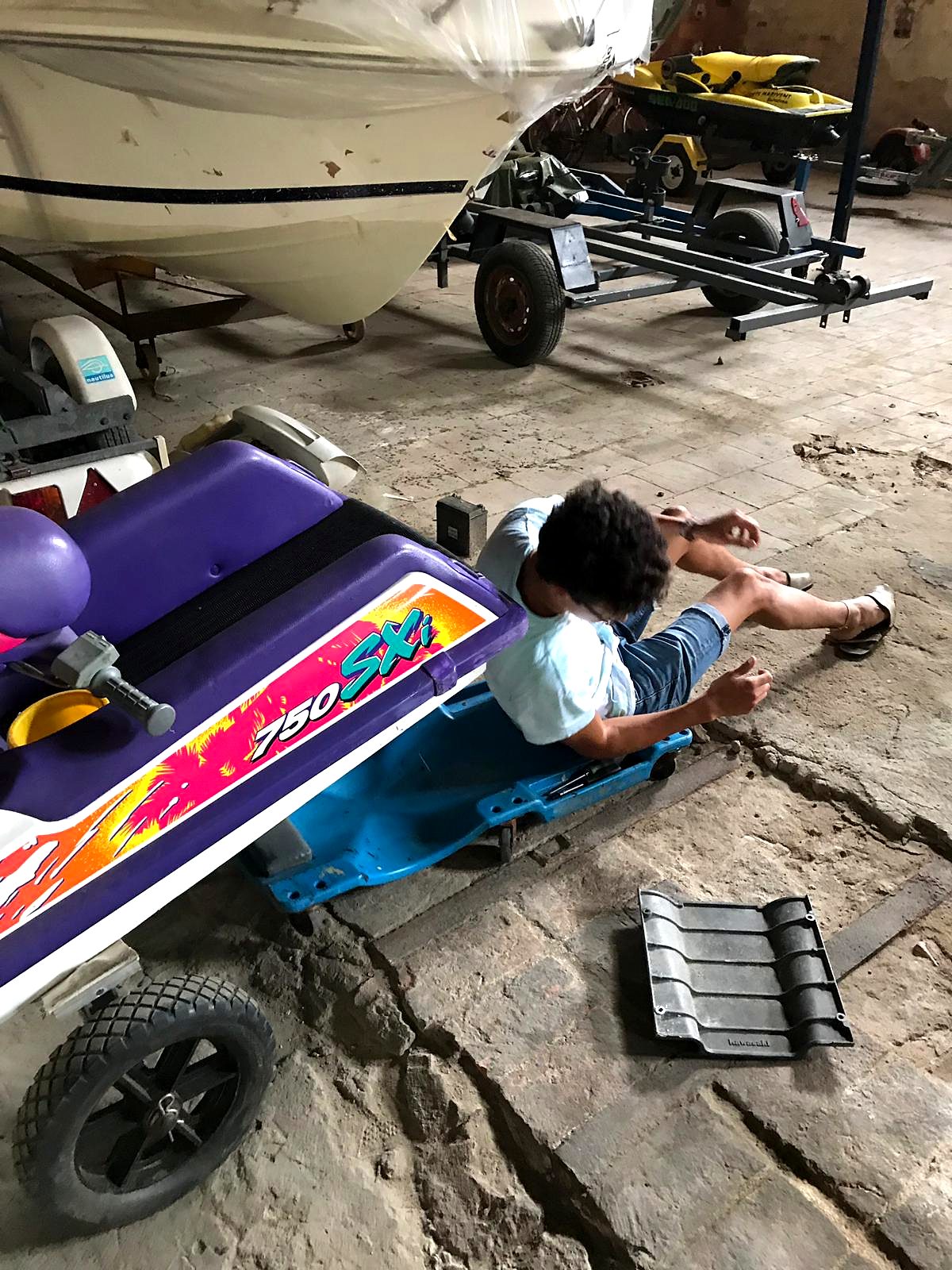 Person on a mechanic creeper next to the purple and white jet ski on a dolly