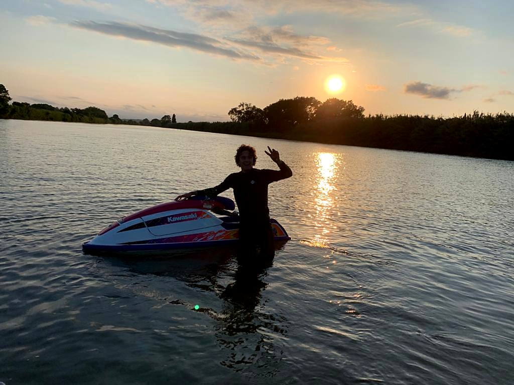 Rai waist-deep in the water at sunset next to a white Kawasaki 750 SXi with pink and purple graphics