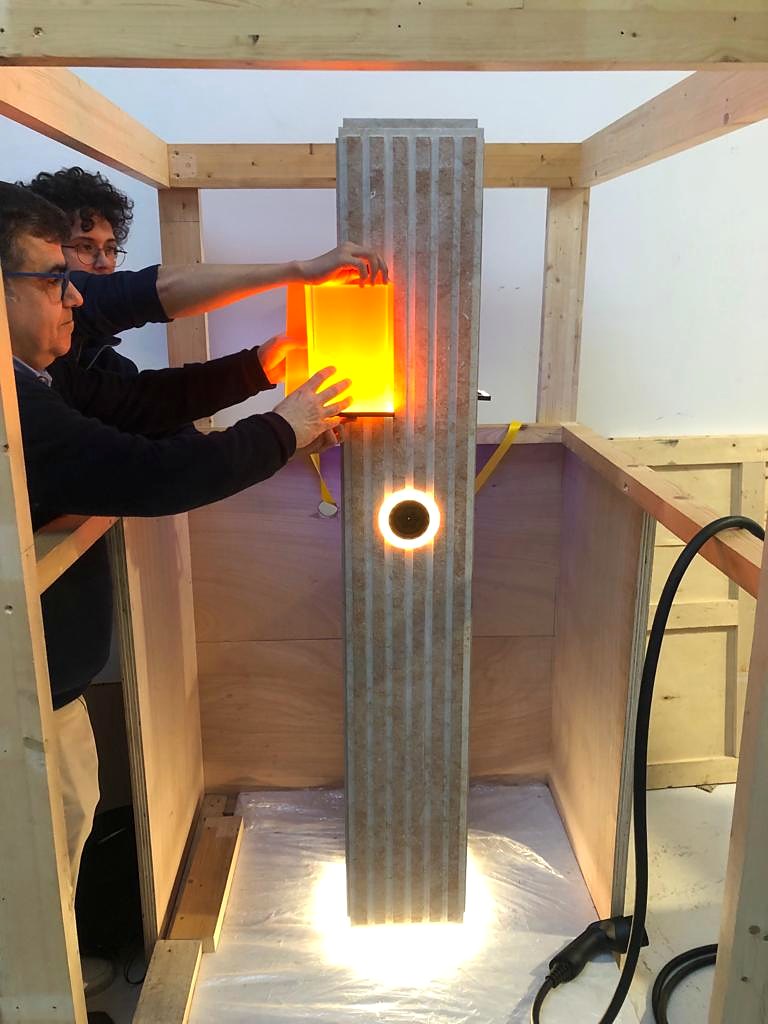 Two people fitting an orange glowing acrylic panel onto a ribbed pillar with a lit ring and base up-light