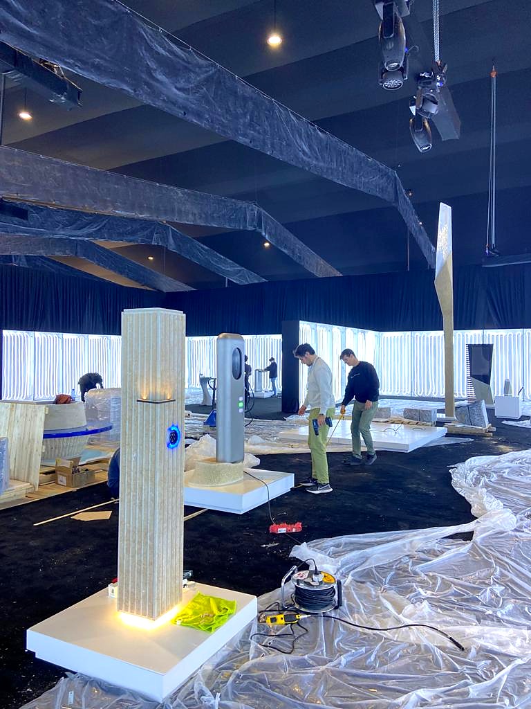 Event hall install: ribbed stone-look pillar with lit charging ring and base light, charger kiosk, faceted light pole, and crew at work